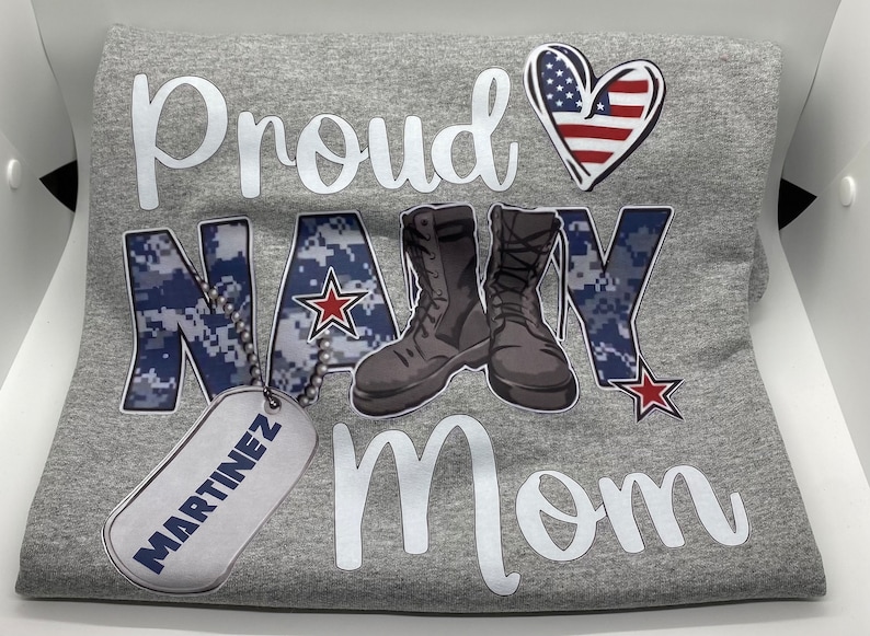 May include: Gray t-shirt with the text "Proud Navy Mom" in white. The word "Navy" is in a blue camouflage pattern with a red star. There are two black boots and a gray dog tag with the text "MARTINEZ" in black.