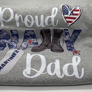 May include: Gray t-shirt with the text "Proud Navy Dad" in white. The word "Navy" is in blue camouflage print with a red star. There are two brown boots and a silver dog tag with the name "Martinez" on it. A red, white, and blue heart is above the text.