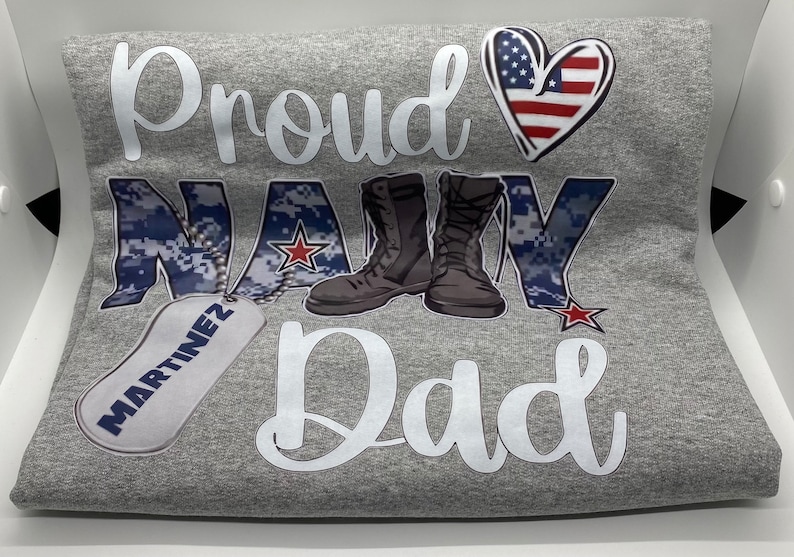 May include: Gray t-shirt with the text "Proud Navy Dad" in white. The word "Navy" is in blue camouflage print with a red star. There are two brown boots and a silver dog tag with the name "Martinez" on it. A red, white, and blue heart is above the text.