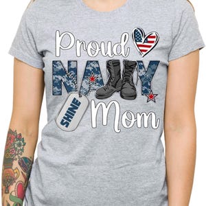 May include: Gray t-shirt with the text "Proud Navy Mom" in white. The text is surrounded by a blue camouflage pattern and a red, white, and blue heart. There is a silver dog tag with the word "Shine" on it.