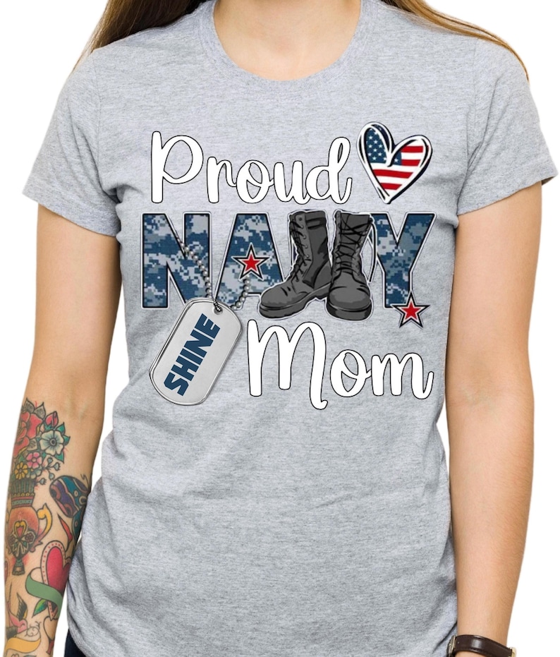 May include: Gray t-shirt with the text "Proud Navy Mom" in white. The text is surrounded by a blue camouflage pattern and a red, white, and blue heart. There is a silver dog tag with the word "Shine" on it.