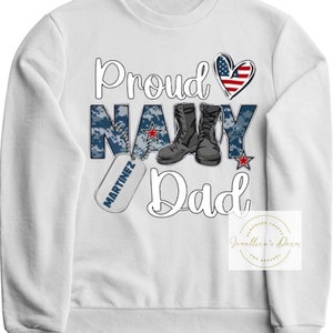 May include: White sweatshirt with the text "Proud Navy Dad" in blue and red. The word "Navy" is in a camouflage pattern. There is a red, white, and blue heart and a military dog tag with the name "Martinez" on it.