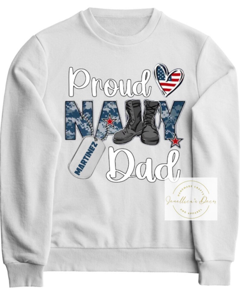 May include: White sweatshirt with the text "Proud Navy Dad" in blue and red. The word "Navy" is in a camouflage pattern. There is a red, white, and blue heart and a military dog tag with the name "Martinez" on it.
