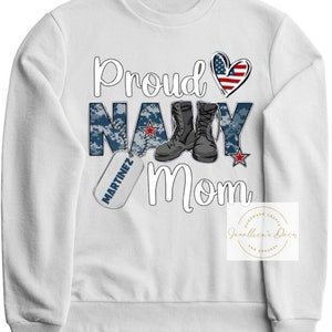 May include: White sweatshirt with the text "Proud Navy Mom" in black. The text is surrounded by a camouflage pattern, a heart with the American flag, a pair of black boots, and a dog tag with the text "MARTINEZ" in black.