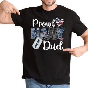 May include: Black t-shirt with a white graphic that says "Proud Navy Dad" with a red heart, an American flag, and a military boot design. The graphic also includes a dog tag with the name "NGUYEN" on it.