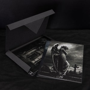 May include: A black box with a flap opening to reveal a set of black and white photographs. The photographs feature a stone angel statue in a cemetery setting.