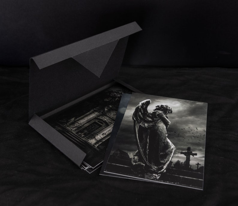 May include: A black box with a flap opening to reveal a set of black and white photographs. The photographs feature a stone angel statue in a cemetery setting.