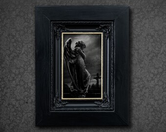 May include: A black framed picture with a gold trim. The picture shows a stone angel statue with wings, standing in a graveyard. The angel is looking down with its head bowed. The background is a dark, cloudy sky.
