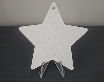 May include: A white star-shaped decorative item with a speckled pattern, displayed on a clear plastic stand. The star has a small hole at the top. The item is on a dark surface.
