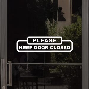May include: A white rectangular vinyl decal applied to a glass door, featuring a rounded border and the bold white text PLEASE KEEP DOOR CLOSED. The design consists of two stacked horizontal sections with rounded corners, clearly visible on the glass.