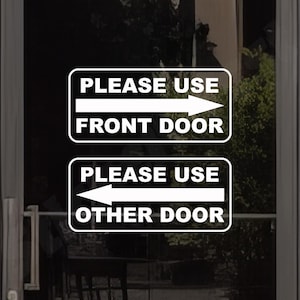 May include: Two rectangular white vinyl decals with rounded corners applied to a glass surface. The top decal features a right-pointing arrow and the text PLEASE USE FRONT DOOR. The bottom decal features a left-pointing arrow and the text PLEASE USE OTHER DOOR.