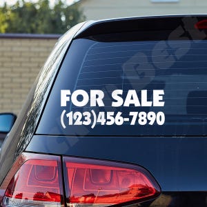 May include: White vinyl decal applied to a car's rear window. The decal reads "FOR SALE" in bold, uppercase letters, with a phone number below. The car is dark blue, and the taillight is visible.