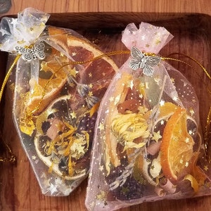 May include: Two small, sheer fabric bags filled with dried citrus fruit, herbs, and gold glitter. The bags are tied with gold ribbon and have a silver butterfly charm.