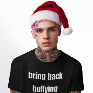以下が含まれることがあります： 黒いTシャツを着て、「bring back bullying」という白い文字が書かれた人。ピンクと白のサンタ帽をかぶっている。