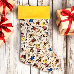 May include: A Christmas stocking with a yellow cuff and a beige body covered in cartoon characters like Snoopy, Charlie Brown, and Woodstock. The stocking is surrounded by wrapped gifts with red ribbons, set against a light wood background.