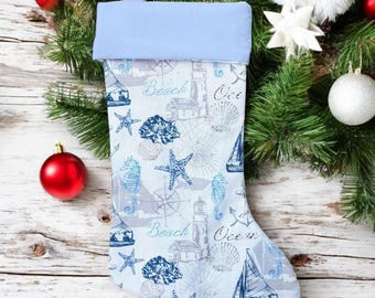May include: A light blue Christmas stocking with a nautical theme. The stocking features a light blue cuff and is decorated with images of sailboats, seashells, starfish, and lighthouses in shades of blue and gray. The words "Beach" and "Ocean" are printed on the stocking.