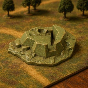 May include: A green, miniature terrain piece resembling a fortress or bunker. The model has a textured, rocky appearance with geometric shapes and a central opening. The background features a green and brown landscape with miniature trees, suggesting a tabletop gaming setting.