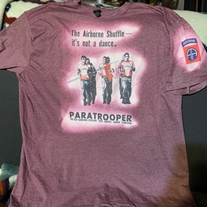 May include: A maroon t-shirt with a faded graphic of four soldiers marching. The graphic includes the text "The Airborne Shuffle... it's not a dance." and "PARATROOPER THE 82D AIRBORNE DIVISION FORT BRAGG, NORTH CAROLINA". The shirt also has a patch on the right sleeve with the text "AIRBORNE" and a stylized "AA" logo.