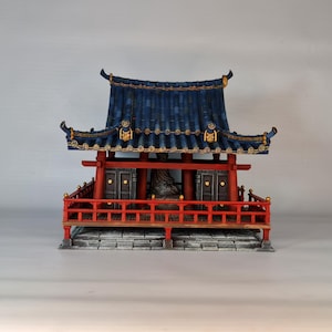 May include: Miniature Asian temple model with a dark blue tiled roof, red supports, and a red railing. The temple has gold accents and two dark doors. The base is gray and the background is a light gray.
