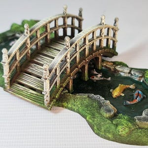 Może przedstawiać: Miniaturowy drewniany mostek pomalowany na brązowo i biało rozciąga się nad małym stawem z zieloną trawą i kamieniami. Staw jest ozdobiony trzema pomarańczowymi i białymi rybami koi.