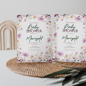 May include: Two floral baby shower invitations with a watercolour design. The invitations are white with a pink, purple, and yellow floral border. The text on the invitations reads "Join us for a Baby Shower in honour of Marigold, Saturday 16th October at two o'clock, 123 Street, Your City, Your Zip, RSVP to [phone number], Registered at Amazon."