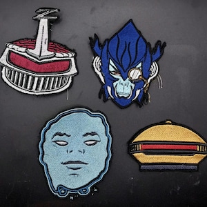 Op de afbeelding: Vier geborduurde patches met unieke ontwerpen. Eén patch heeft een rood en wit helmachtig ontwerp. Een andere toont een blauw aapachtig gezicht met een monocle. Een derde patch is een lichtblauw gezicht. De laatste is geel en zwart.