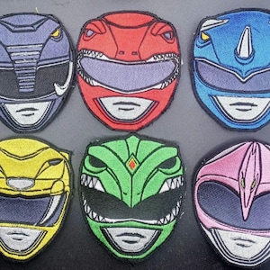 以下が含まれることがあります： 紫、赤、青、黄、緑、ピンクの様々な色のパワーレンジャーのヘルメットが刺繍された6つのパッチ。各パッチは、詳細なステッチと独特の顔のデザインを示しています。
