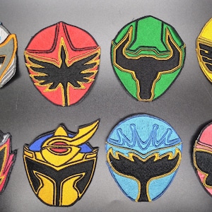 Puede incluir: Una colección de ocho parches bordados con diseños de cascos estilizados y coloridos. Cada parche tiene una forma y una combinación de colores únicas, incluyendo rojo, verde, amarillo, azul y rosa, con detalles en negro y dorado. Los parches son probablemente para disfraces o manualidades.