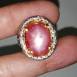 Puede incluir: Un anillo con una gran piedra preciosa rosa de forma ovalada rodeada por un halo de piedras blancas más pequeñas. El anillo está engastado en plata y tiene una banda de oro.