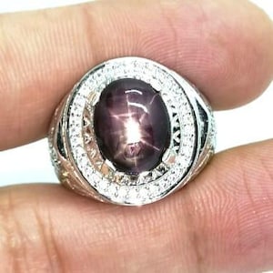 Puede incluir: Un anillo de plata con una gran gema ovalada en forma de estrella. La gema es de color púrpura oscuro con un patrón de estrella de seis puntas. El anillo está rodeado por un halo de pequeñas piedras transparentes. El anillo está sostenido por los dedos de una persona.