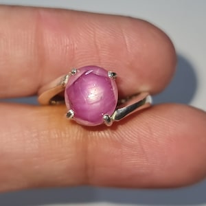 May include: A silver ring featuring a pink star ruby gemstone. The ruby is set in a prong setting and displays a star-shaped pattern.