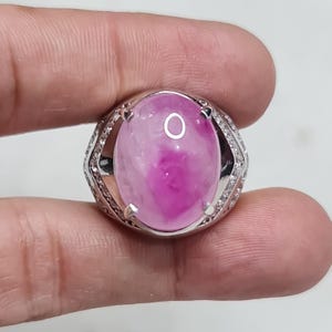 Könnte beinhalten: Ein silberner Ring mit einem großen, ovalen rosa Edelstein. Der Edelstein hat einen helleren Rosaton mit einem dunkleren rosa Zentrum. Das Ringband ist silberfarben und hat kleine, klare Steine um den Edelstein.