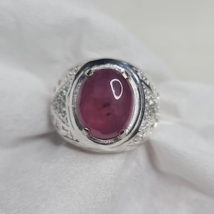 Peut inclure: Une bague en argent ornée d'une pierre précieuse ovale rouge foncé. La bague a un anneau détaillé et texturé avec de petites pierres d'accent claires. La pierre précieuse est le point central, présentant une couleur riche et saturée et une surface polie.