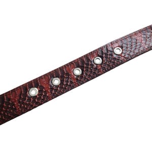 Może przedstawiać: Zbliżenie brązowo-czerwonego paska ze wzorem sk&oacute;ry węża i srebrnymi oczkami. Pasek wykonany jest z teksturowanego materiału i posiada kilka r&oacute;wnomiernie rozmieszczonych otwor&oacute;w do regulacji. Pasek jest ustawiony po przekątnej.