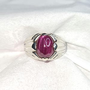 May include: A silver-toned ring featuring a central oval-shaped cabochon star ruby with a distinct six-rayed star effect. The metal band displays a polished finish with decorative grooved detailing extending from the stone setting.