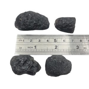 Può includere: Quattro rocce scure e strutturate di varie dimensioni sono esposte accanto a un righello. Il righello mostra sia pollici che centimetri, con la roccia più grande che misura circa 5 cm di lunghezza.