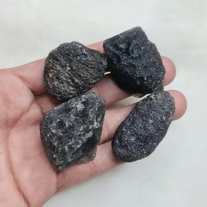 Puede incluir: Se muestran cuatro piedras de color gris oscuro y textura rugosa de varias formas. Las piedras se sostienen en una mano, mostrando su apariencia natural y sin pulir. Las piedras tienen un color gris oscuro con algunas áreas grises y marrones más claras.