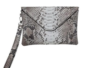 May include: A rectangular clutch bag featuring a grey and white snakeskin pattern with a triangular envelope-style flap closure. The bag includes a matching detachable wrist strap attached to a metal clip on the side, showcasing a textured finish.