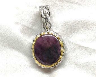 May include: A round pendant featuring a dark purple, opaque cabochon stone set in a gold-toned bezel. The stone is surrounded by a scalloped silver-toned frame with a textured, organic-looking bail at the top for attaching to a chain.