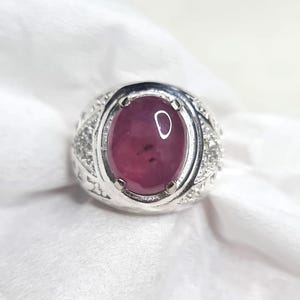 Puede incluir: Un anillo de plata con una piedra preciosa ovalada de color rojo intenso. La banda del anillo tiene un diseño detallado y texturizado con pequeños acentos brillantes. La piedra preciosa es el punto focal del anillo.