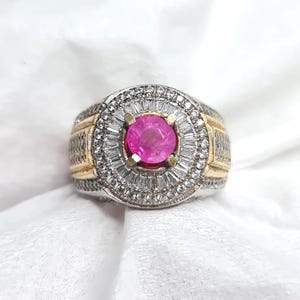 May include: A silver and gold ring with a large, round, bright pink gemstone at the center. The gemstone is surrounded by smaller, clear, rectangular stones. The ring has a detailed design on the band.