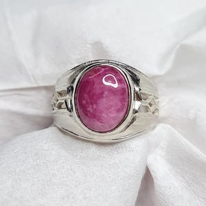 May include: A silver ring featuring a large, oval-shaped, pink gemstone. The ring has a wide band with decorative cutouts on the sides. The gemstone is a vibrant shade of pink, set in a polished silver setting.