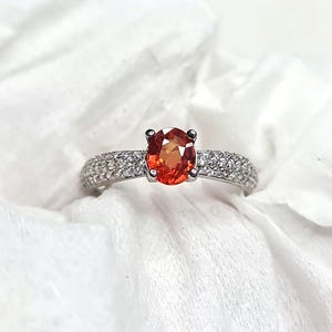 May include: A silver ring featuring a large, oval-cut orange gemstone as the centerpiece. The band is encrusted with small, clear gemstones. The ring is set against a white, textured background, highlighting the jewelry's sparkle.