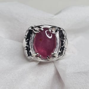 May include: A silver ring featuring a large, oval-shaped, deep red gemstone. The ring has an ornate, detailed setting with dark accents. The gemstone is the focal point, set against a white background.
