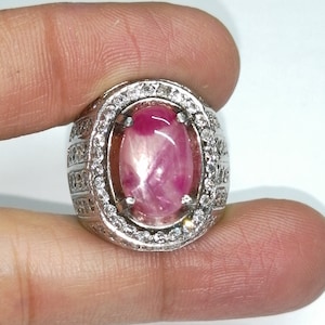 May include: A silver ring featuring a large oval pink ruby gemstone, encircled by smaller white gemstones.