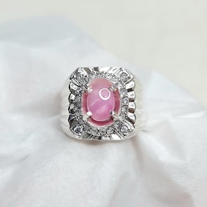 Könnte beinhalten: Ein silberner Ring mit einem großen, oval geschliffenen rosa Edelstein in der Mitte, umgeben von kleineren, klaren Steinen. Der Ring hat ein detailliertes, strukturiertes Band und eine quadratische Fassung, die einen klassischen und eleganten Look erzeugt. Der Ring liegt auf einer weißen Oberfläche.