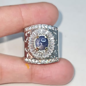 Op de afbeelding: Een zilveren ring met een centrale ovale blauwe edelsteen, omringd door kleinere, heldere stenen. De ring heeft een rechthoekige band met decoratieve details. De edelsteen heeft een stervormig patroon. De ring wordt tussen vingers gehouden.