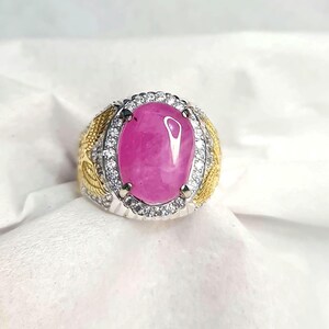 May include: A ring featuring a large, oval-shaped, smooth pink cabochon stone set in a silver-toned metal frame. The center stone is surrounded by a halo of small, clear, sparkling stones, with gold-toned wing-like accents on the sides.