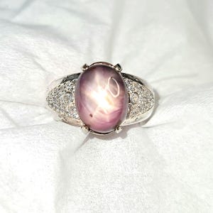 Peut inclure: Une bague en argent avec une grande pierre précieuse ovale de couleur violette taillée en cabochon avec un effet étoile. L'anneau est orné de nombreuses petites pierres rondes transparentes en pavé et de deux pierres plus grandes sur les côtés.