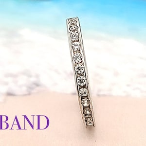 May include: A thin silver band with a row of small clear cubic zirconia stones set around the entire band. The band is photographed against a blurred background of a beach with white sand and blue water. The text "CZ BAND" is visible in the lower left corner of the image.
